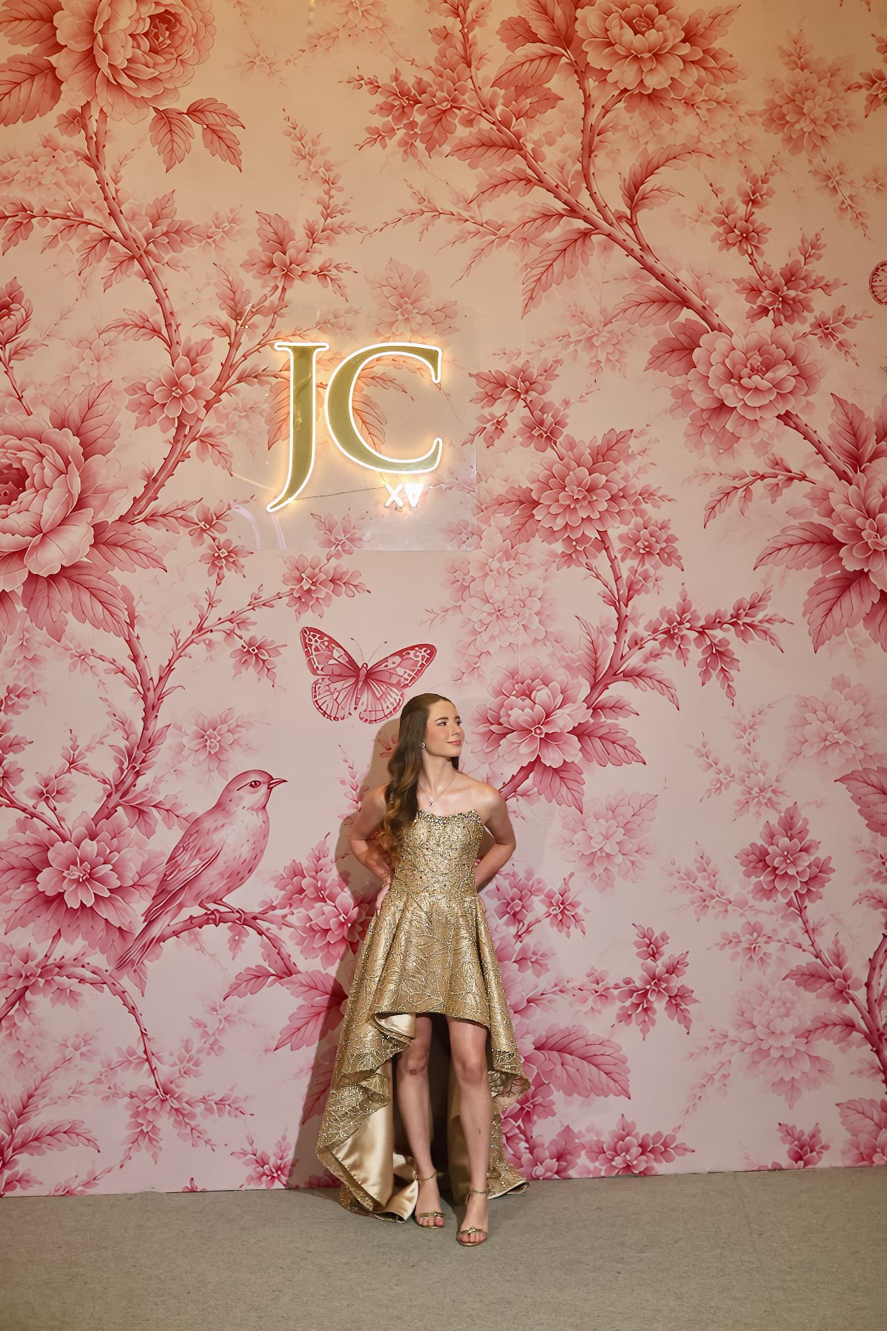 Debutante em vestido dourado diante de parede floral com monograma luminoso da festa de quinze anos